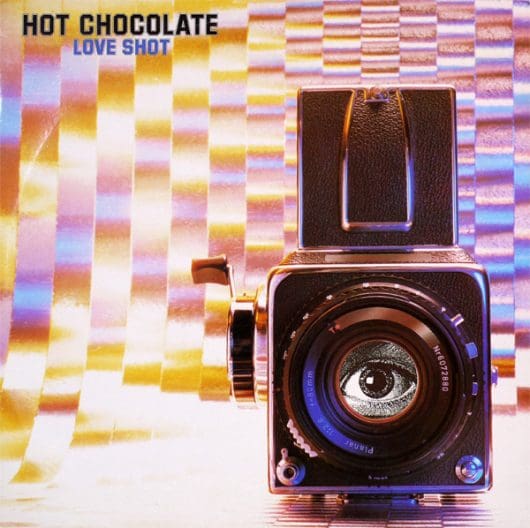 An image of Hot Chocolate's studio album Love Shot. This album cover features an old analogue camera with a black and white image of an eye where the lens should be. The background is a shiny, psychedelic print with lettering in the top left-hand corner reading 'Hot Chocolate' in black block letters and 'Love Shot' in identical smaller violet letters underneath.