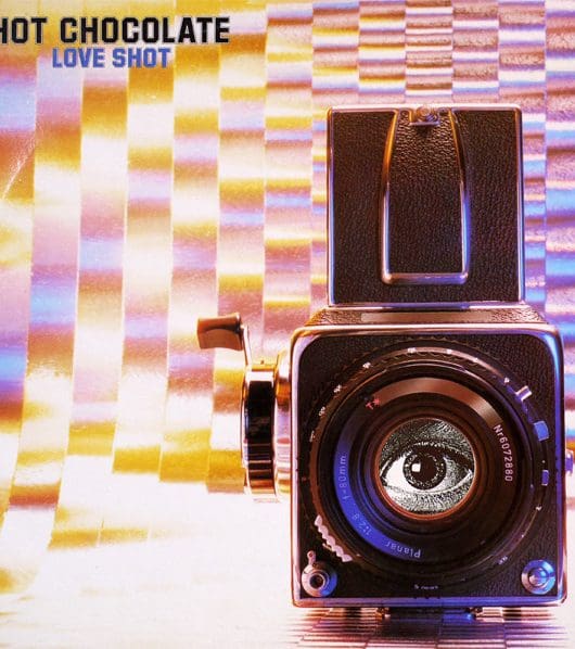 An image of Hot Chocolate's studio album Love Shot. This album cover features an old analogue camera with a black and white image of an eye where the lens should be. The background is a shiny, psychedelic print with lettering in the top left-hand corner reading 'Hot Chocolate' in black block letters and 'Love Shot' in identical smaller violet letters underneath.